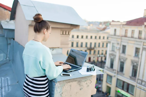 Schöne und stilvolle Mädchen arbeitet für einen Laptop auf dem Dach des Hauses in der Altstadt. Auf dem Tisch liegen auch Dokumente und eine alte Kamera — Stockfoto