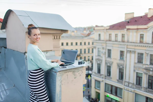 Güzel ve şık bir kız eski şehir Evin çatısı bir laptop için çalışır. Masanın üzerine de belgeleri ve eski fotoğraf makinesi için