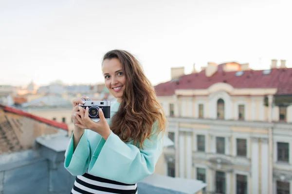 Look de moda, modelo jovem muito legal com câmera de filme retro. cabelo encaracolado ao ar livre. Fotógrafo menina elegante leva a cidade velha do telhado ao pôr do sol — Fotografia de Stock