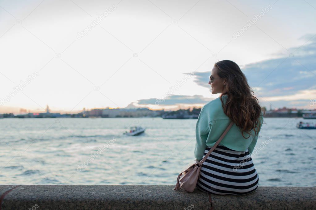 Young and beautiful girl sitting on the embankment of the river. she looks at the sunset and ships passing by. Saint Petersburg, Russia