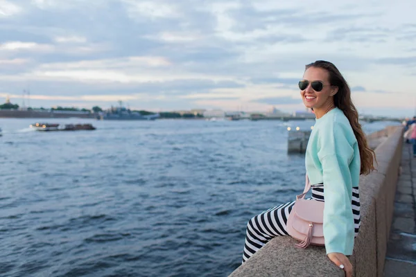 Menina jovem e bonita sentada no dique do rio. Ela olha para o pôr-do-sol e para os navios que passam. São Petersburgo, Rússia — Fotografia de Stock