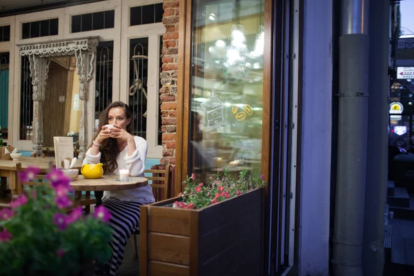 Молода приваблива дівчина сидить увечері в яскравому і теплому кафе з чашкою чаю і дивиться у вікно. Вулиця темна і холодна. Концепція комфорту — стокове фото