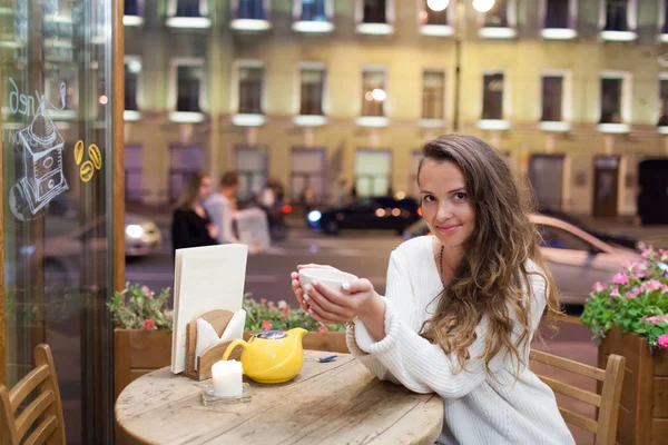 Menina atraente jovem sentado à noite em um café com uma xícara de chá para o pano de fundo de carros que passam e vida na cidade. Ela olha para a câmera e sorri — Fotografia de Stock