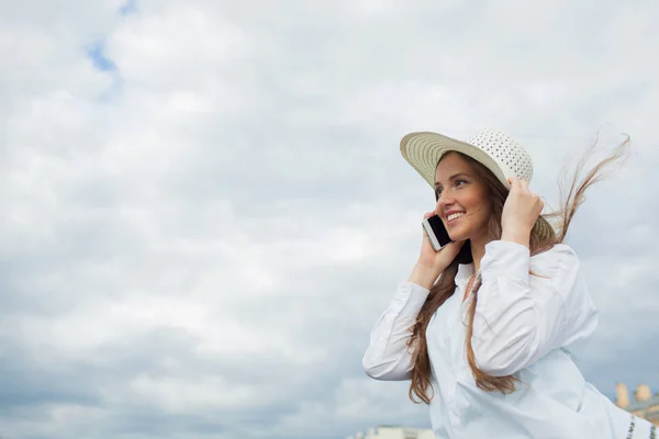 Uma menina bonita e sorridente em um chapéu branco com aba larga está de pé na ponte e falando ao telefone contra o fundo de nuvens azuis — Fotografia de Stock