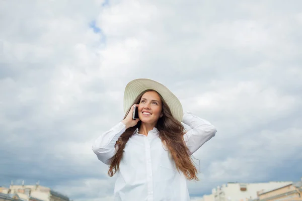 Uma menina bonita e sorridente em um chapéu branco com aba larga está de pé na ponte e falando ao telefone contra o fundo de nuvens de tempestade azul — Fotografia de Stock