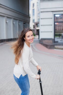 Yeni bir konut kompleksi içinde bir scooter sürme bir ışık kazaklı uzun kahverengi saçlı genç ve çekici kız
