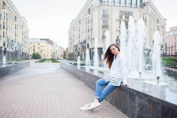 Young and beautiful girl sitting at the fountain and smiling in a new residential complex — Stock Photo, Image