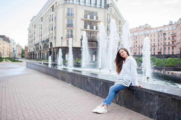 Młoda i piękna dziewczyna siedzi przy fontannie i uśmiechając się w nowym kompleksie mieszkaniowym — Zdjęcie stockowe