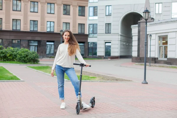 Een jong en aantrekkelijk meisje met lang bruin haar in een lichte trui rijden een scooter in een nieuw wooncomplex — Stockfoto