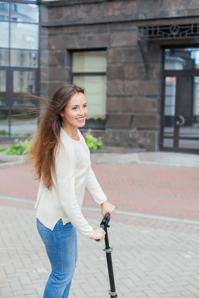 Una ragazza giovane e attraente con lunghi capelli castani in un maglione leggero in sella a uno scooter in un nuovo complesso residenziale — Foto Stock