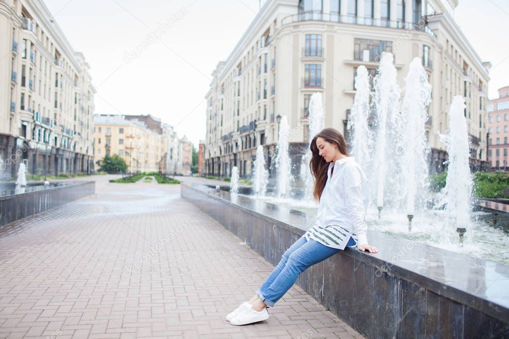 Young and beautiful girl sitting at the fountain and smiling in a new residential complex