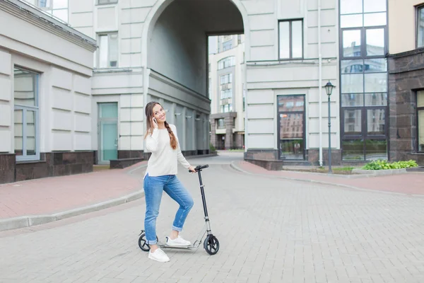 Uzun kahverengi saçlı güzel genç kız arkadaşınıza arka planda yeni yerleşim bölgesi telefonda konuşmak için scooter sürme sırasında durdu. Kentsel arka plan