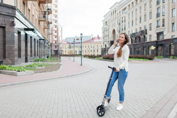 Uzun kahverengi saçlı güzel genç kız arkadaşınıza arka planda yeni yerleşim bölgesi telefonda konuşmak için scooter sürme sırasında durdu. Kentsel arka plan