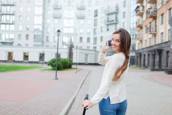 Hermosa joven con el pelo castaño largo se detuvo mientras montaba en el scooter para hablar con un amigo por teléfono en el fondo del nuevo barrio residencial. Fondo urbano — Foto de Stock