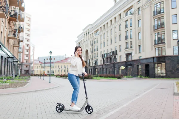 Beautiful young girl with long brown hair stopped while riding the scooter to talk to a friend on the phone on the background of the new residential quarter. Urban background — Stock Photo, Image