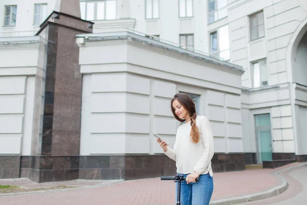 Hermosa, joven y de dientes blancos chica con el pelo largo y castaño se detuvo mientras montaba en la moto, para escribir a un amigo por teléfono. Ella está vestida con un suéter blanco y vaqueros azules. Fondo urbano —  Fotos de Stock