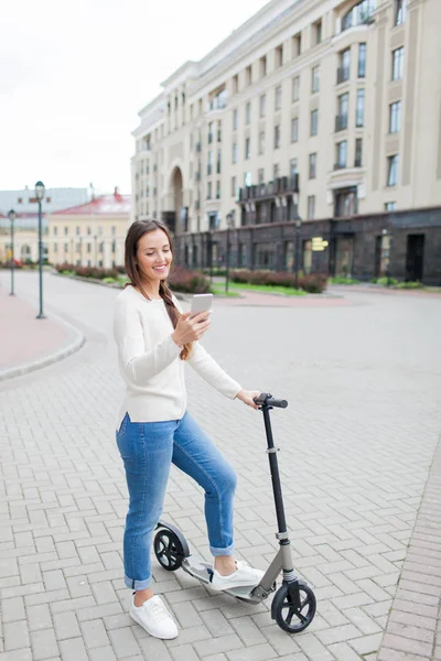 Beautiful, young, and white-toothed girl with long brown hair stopped while riding the scooter, to write to a friend on the phone. She is dressed in a white sweater and blue jeans. Urban background — Stock Photo, Image