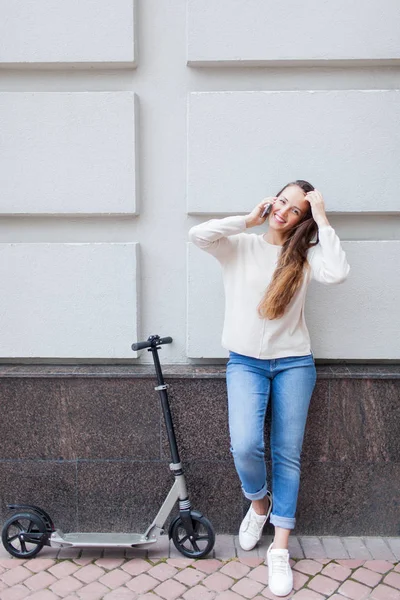 Rapariga bonita com cabelo castanho longo parou ao andar de scooter, para chamar um amigo no telefone no fundo da parede cinza. Ela está vestida com uma camisola branca e jeans azul — Fotografia de Stock