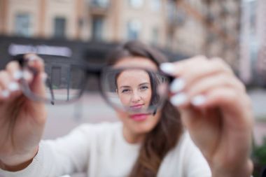 Closeup portrait of young women with glasses. She has eyesight problems and is squinting his eyes a little bit. Beautiful girl is holding eyeglasses right in front of camera with two hand clipart