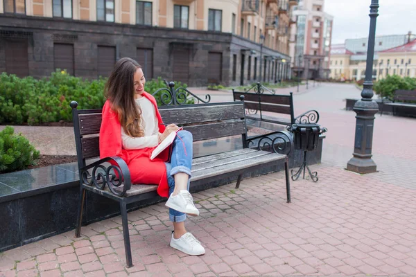 Uma garota atraente com cabelos castanhos longos e um sorriso de dente branco se senta em um banco e escreve seus pensamentos sobre o fundo urbano em um caderno vermelho. Ela está vestindo uma camisola branca e jeans azul — Fotografia de Stock