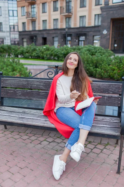 Una chica atractiva se sienta en un banco y escribe sus pensamientos sobre el fondo urbano en un cuaderno rojo. Lleva un suéter blanco, vaqueros azules y una tela a cuadros roja. Una mujer está pensando en una idea —  Fotos de Stock