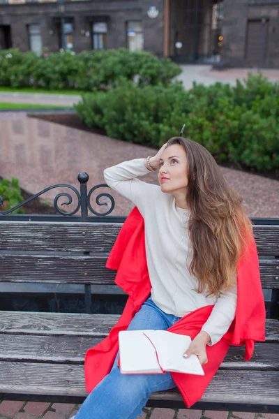 Sitzt ein attraktives Mädchen auf einer Bank und schreibt ihre Gedanken über den urbanen Hintergrund in ein rotes Notizbuch. Sie trägt einen weißen Pullover, blaue Jeans und einen roten Karo. eine Frau denkt über eine Idee nach — Stockfoto