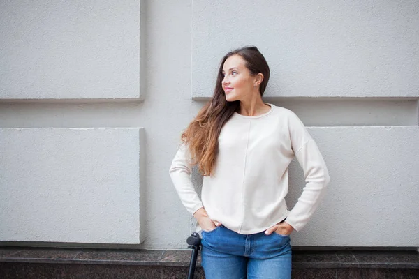 Rapariga bonita com cabelo castanho longo parou enquanto andava de scooter no fundo da parede cinza. Ela está vestida com uma camisola branca e jeans azul — Fotografia de Stock