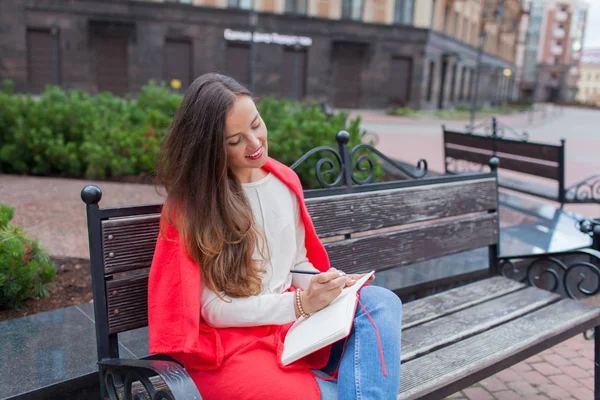 Una chica atractiva con el pelo castaño largo y una sonrisa de diente blanco se sienta en un banco y escribe sus pensamientos sobre el fondo urbano en un cuaderno rojo. Lleva un suéter blanco y vaqueros azules. — Foto de Stock