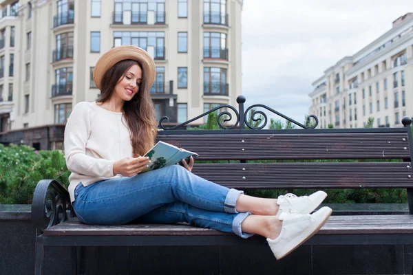 Una joven hermosa mujer con un sombrero elegante se sienta en un banco en un nuevo barrio residencial y lee un libro de papel. Ella hojea las páginas y sonríe — Foto de Stock