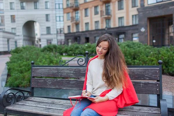 Una chica atractiva con el pelo castaño largo se sienta en un banco y escribe sus pensamientos sobre el fondo de la ciudad en un cuaderno rojo. Lleva un suéter blanco, vaqueros azules y un cuadros rojos. —  Fotos de Stock