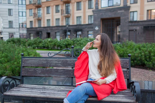 An attractive girl with long brown hair sits on a bench, hiding behind a red rug, gnawing a pen and thinking on an urban background. She has a notebook in her hands — Stock Photo, Image