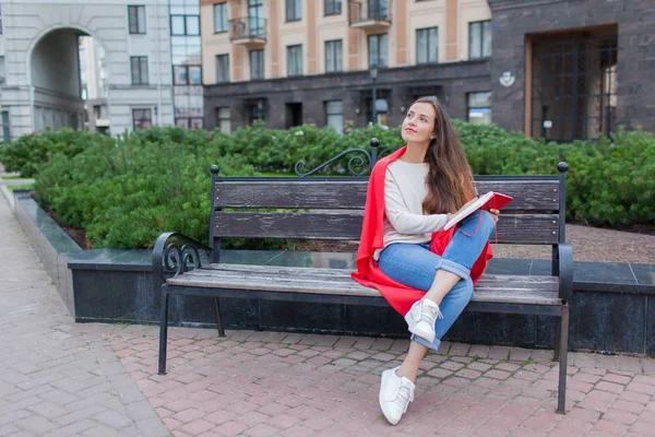 An attractive girl sits on a bench and writes her thoughts on the urban background in a red notebook. She wears a white sweater, blue jeans and a red plaid. A woman is thinking over an idea — Stock Photo, Image