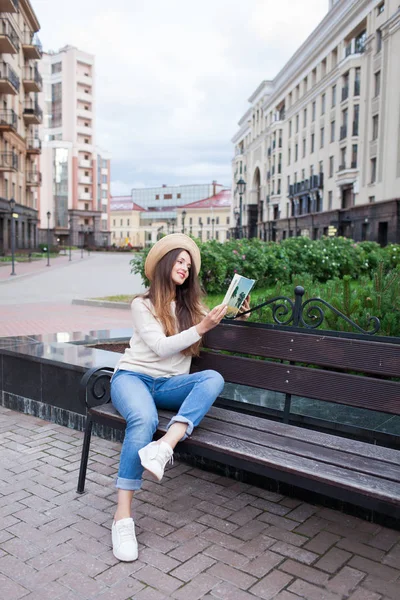 Una joven hermosa mujer con un sombrero elegante se sienta en un banco en un nuevo barrio residencial y lee un libro de papel. Ella hojea las páginas y sonríe. Fondo urbano —  Fotos de Stock