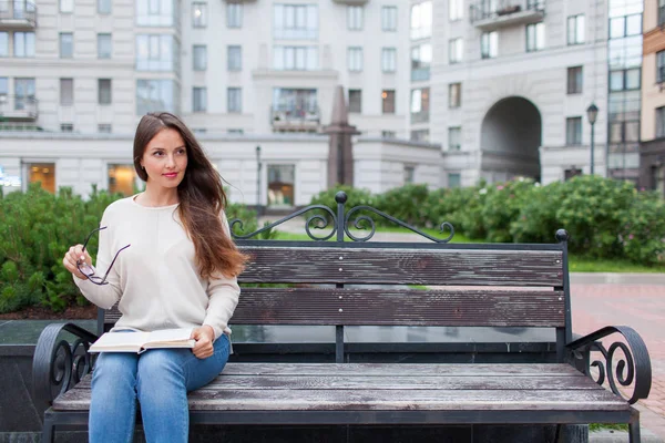 Una hermosa joven con el pelo castaño largo sentada en el banco con el libro y las gafas en las manos. Salió de la casa por la noche para leer en el patio. — Foto de Stock