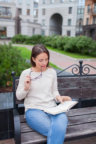 Una bella ragazza con lunghi capelli castani seduta sulla panchina con libro e bicchieri da morso durante la lettura. Uscì di casa in una calda serata per leggere in cortile. Lo sfondo urbano — Foto Stock