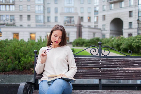 Ein schönes junges Mädchen mit langen braunen Haaren sitzt auf der Bank mit Buch und beißender Brille beim Lesen. Sie verließ das Haus an einem warmen Abend, um im Hof zu lesen. der städtische Hintergrund — Stockfoto