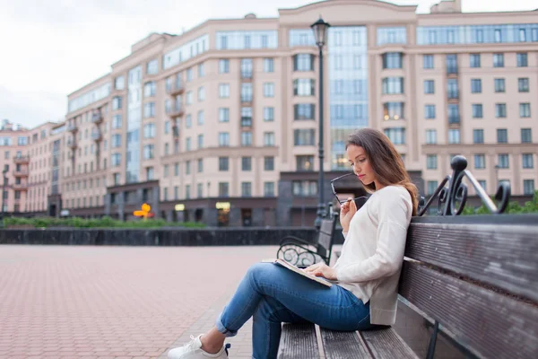 Una hermosa joven con el pelo castaño largo sentada en el banco con libro y gafas mordedoras mientras lee. Salió de la casa en una cálida noche para leer en el patio. El contexto urbano — Foto de Stock