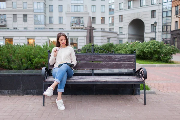 A beautiful young girl with long brown hair sitting on the bench with book and biting glasses while reading. She left the house on a warm evening to read in the yard. The urban background — Stock Photo, Image