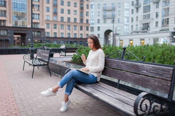 Ein schönes junges Mädchen mit langen braunen Haaren, das mit einem Buch auf einer Bank sitzt und eine Brille hält. Sie verließ das Haus an einem warmen Abend, um im Hof zu lesen. der städtische Hintergrund. — Stockfoto
