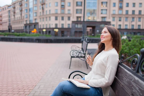 Ein schönes junges Mädchen mit langen braunen Haaren, das auf der Bank sitzt und das Buch und die Brille in den Händen hält. Abends verließ sie das Haus, um im Hof zu lesen. Urbaner Hintergrund — Stockfoto