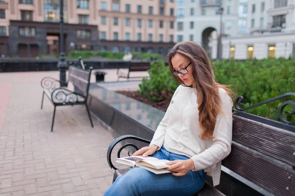 Junge schöne Mädchen mit Brille lange braune Haare sitzt auf einer Bank mit einem Buch. Sie verließ das Haus an einem warmen Abend, um im Hof zu lesen. der städtische Hintergrund. — Stockfoto