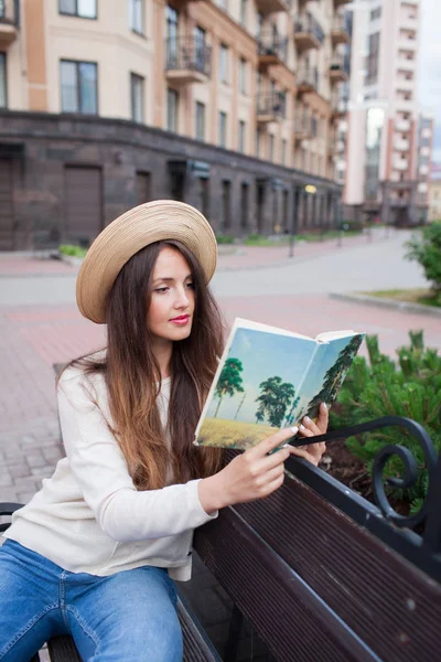 En ung vacker kvinna i en elegant hatt sitter på en bänk i ett nytt bostadsområde och läser en bok i papper. Hon vänder igenom sidorna. Urban bakgrund — Stockfoto