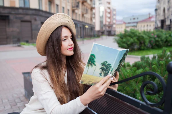 En ung vacker kvinna i en elegant hatt sitter på en bänk i ett nytt bostadsområde och läser en bok i papper. Hon vänder igenom sidorna. Urban bakgrund — Stockfoto