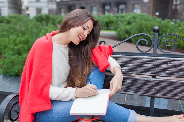 Attraktives Mädchen, das mit nackten Füßen und einer roten Decke auf einer Bank im neuen Wohngebiet sitzt und seine Gedanken in ein rotes Notizbuch schreibt. der städtische Hintergrund — Stockfoto