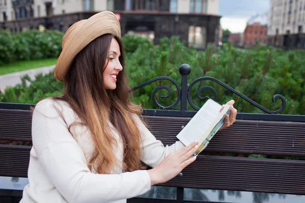 Una joven hermosa mujer con un sombrero elegante se sienta en un banco en un nuevo barrio residencial y lee un libro de papel. Ella hojea las páginas y sonríe. Fondo urbano —  Fotos de Stock