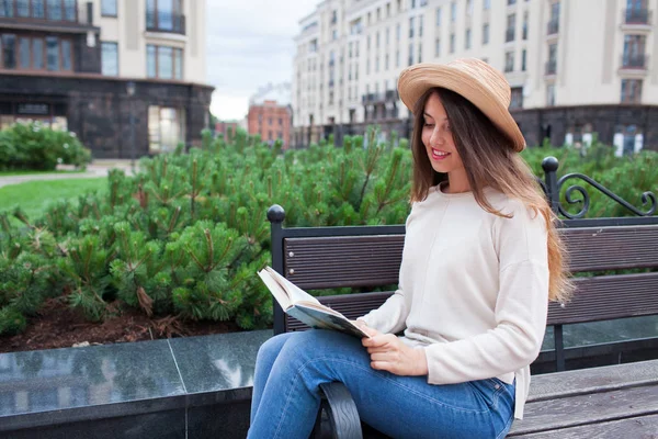 Una joven hermosa mujer con un sombrero elegante se sienta en un banco en un nuevo barrio residencial y lee un libro de papel. Ella hojea las páginas y sonríe. Fondo urbano —  Fotos de Stock