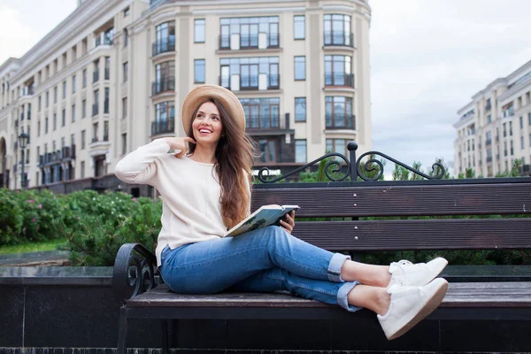 Uma mulher bonita nova em um chapéu elegante senta-se em um banco em um bairro residencial novo e lê um livro de papel. Ela vira as páginas e sorri. Fundo urbano — Fotografia de Stock