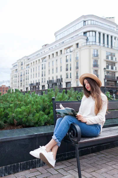 Uma mulher bonita nova em um chapéu elegante senta-se em um banco em um bairro residencial novo e lê um livro de papel. Ela vira as páginas. Fundo urbano — Fotografia de Stock