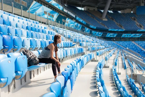 Sexy girl posing at stadium with the backpack. Fitness girl with a sports figure in black leggings sitting on the seat in the stadium. Sport woman posing at stadium. Girl smiling after sport day