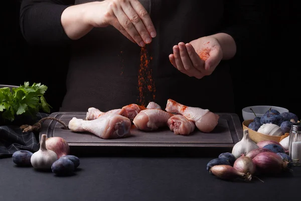 Una chef espolvorea palitos de pollo crudos frescos sobre un fondo oscuro. Cerca se encuentran los ingredientes para cocinar: chalotas, ciruelas azules, ajos, sal y perejil — Foto de Stock